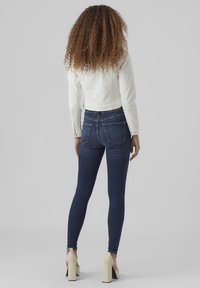 Un jean skinny bleu foncé à taille haute, associé à une veste en denim blanche cropped. Des cheveux bouclés et des chaussures à talons hauts beiges sont visibles.