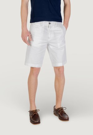 Shorts blancs avec une texture lisse, présentant des poches avant et une fermeture à boutons. Associés à des chaussures à lacets marron et un haut bleu marine.