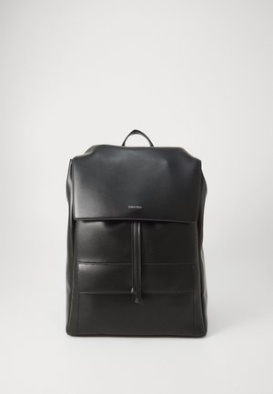 Zaino in pelle nera con parte superiore ripiegata, tasca anteriore con zip e cuciture a vista. Presenta una maniglia e un logo minimalista. Texture liscia.