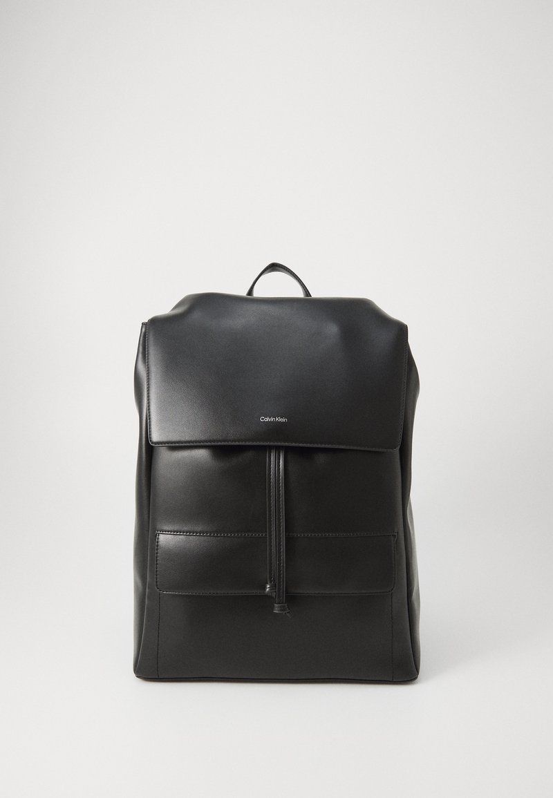 Zaino in pelle nera con parte superiore ripiegata, tasca anteriore con zip e cuciture a vista. Presenta una maniglia e un logo minimalista. Texture liscia.