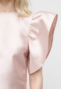 Γυναίκα που φοράει ένα ανοιχτό ροζ σατέν τοπ με ένα μεγάλο, δομημένο, φουσκωτό μανίκι στο δεξί της χέρι.