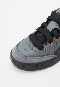 Zapatilla gris con puntera y cordones gruesos negros, ojales naranjas y parte superior de cuero perforado sobre una suela negra, todo contra un fondo blanco.