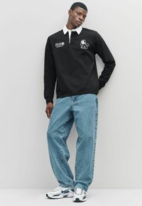 Polo negra de manga larga con cuello blanco, que presenta un logotipo de gato de dibujos animados y el texto "FELIX", combinada con jeans de mezclilla azules claros y zapatillas blancas.
