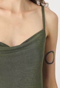 Olivu zaļš, pieguļošs tank top ar plānām siksnām, ar V veida dekoltē un mīkstu, teksturētu audumu. Redzams āda dekoltē rajonā.