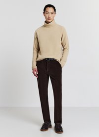 Un pull à col roulé en tricot chunky beige associé à un pantalon en velours côtelé marron foncé et des chaussures à lacets marron, se tenant devant un fond uni.