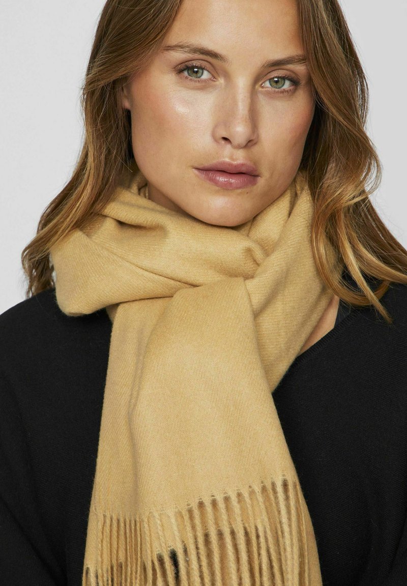 Écharpe couleur camel en matériau doux, avec des extrémités frangées, drapée de manière élégante, au toucher lisse et à un éclat subtil.