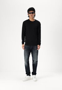 Schwarzer Pullover mit langen Ärmeln, dunkelblaue Jeans und weiße Sneakers. Die Accessoires umfassen schwarze Sonnenbrillen; das Modell steht vor einem schlichten Hintergrund.