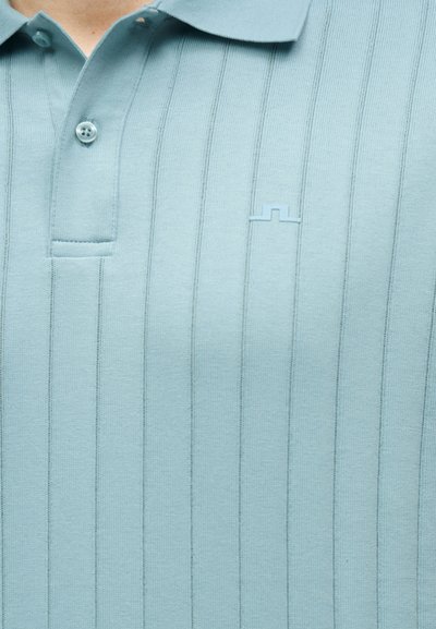 Ανοιχτό μπλε ριμπ πουκάμισο πόλο με κάθετες ρίγες και μικρό ανυψωμένο λογότυπο στο αριστερό στήθος, κουμπωμένο κοντά στον γιακά.