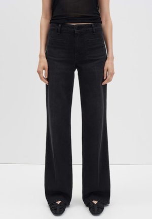 Vrouw in hoge zwarte wijde jeans, zwart tanktop en zwarte puntige schoenen die tegen een witte achtergrond staat.