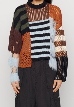 Woman wearing a multicolored, patchwork knit sweater with stripes, mesh, fuzzy textures, and various knit patterns paired with loose black pants.