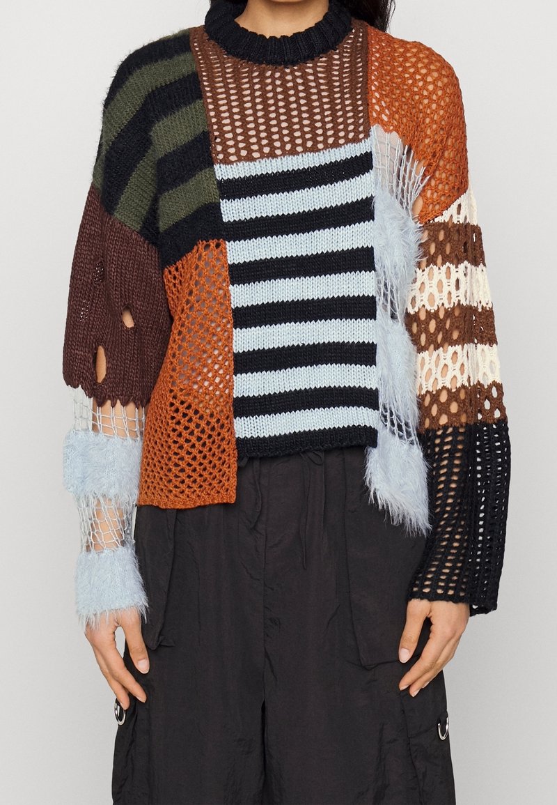 Femme portant un pull en tricot patchwork multicolore avec des rayures, du mesh, des textures duveteuses et divers motifs de tricot, associé à un pantalon noir ample.