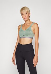 Sujetador deportivo verde con escote en V, tejido texturizado, tirantes anchos y un patrón de malla decorativo. Combinado con leggings negros de cintura alta.