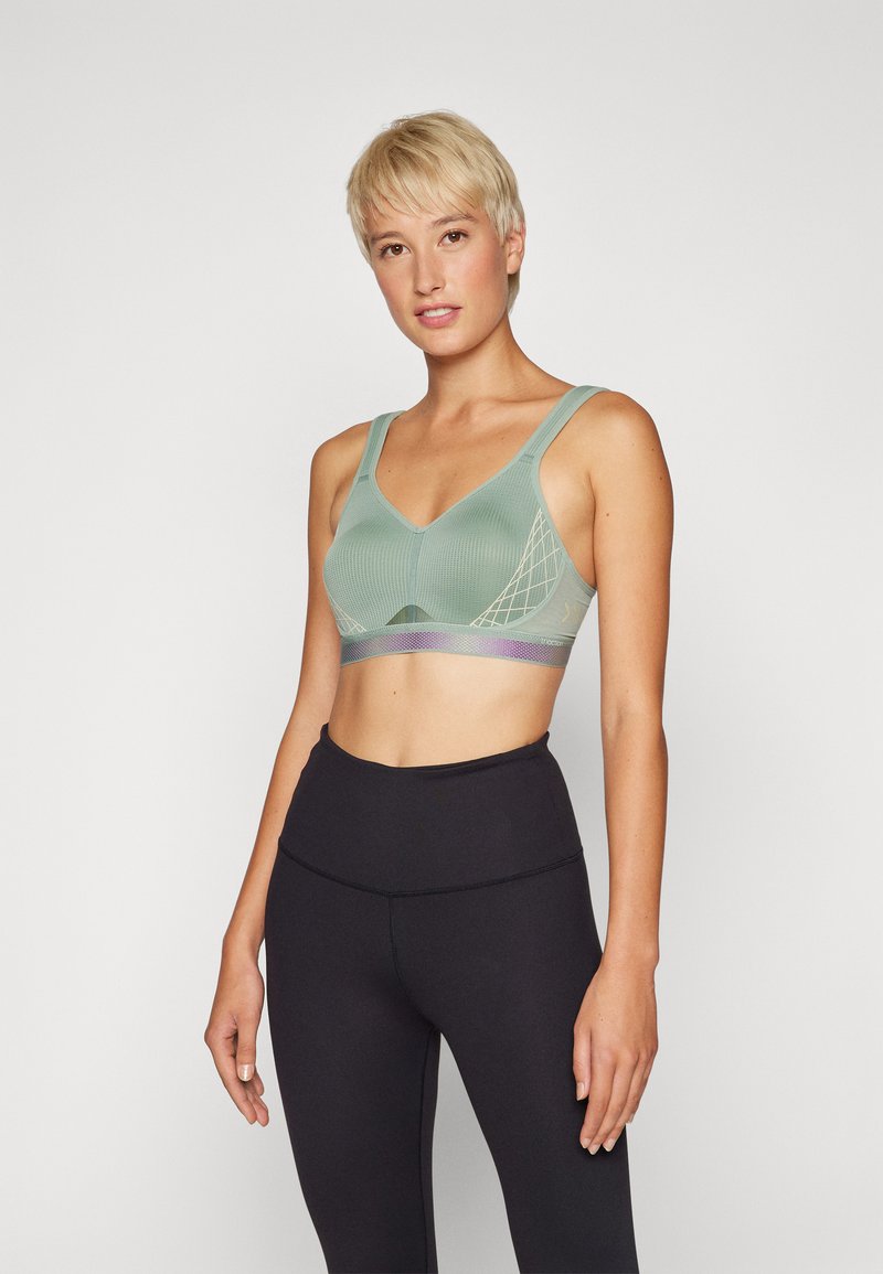 Sujetador deportivo verde con escote en V, tejido texturizado, tirantes anchos y un patrón de malla decorativo. Combinado con leggings negros de cintura alta.