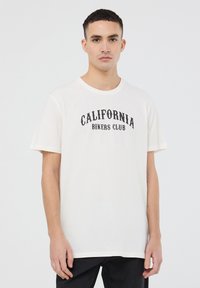 T-shirt beige chiaro con maniche corte, caratterizzato da testo nero "CALIFORNIA BIKERS CLUB" sulla parte anteriore. Tessuto in cotone morbido, vestibilità comoda.