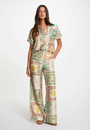 Donna che indossa una camicia a maniche corte ampie e pantaloni a gamba larga con un motivo geometrico e floreale multicolore, in piedi con una mano in tasca.