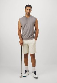 Top sin mangas gris claro, pantalones cortos crema, sosteniendo un palo de golf. Zapatos blancos con detalles en azul marino y calcetines blancos. Diseño minimalista.