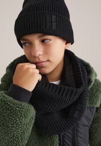 Fant s črno pleteno kapo in ujemajočim šalom, oblečen v zeleni flisjakni, ki gleda vstran z roko blizu obraza.