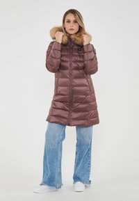 Donna che indossa un lungo giubbotto imbottito color borgogna con cappuccio in pelliccia, mentre tiene il cappuccio, abbinato a jeans a gamba larga di colore azzurro chiaro e sneakers bianche.