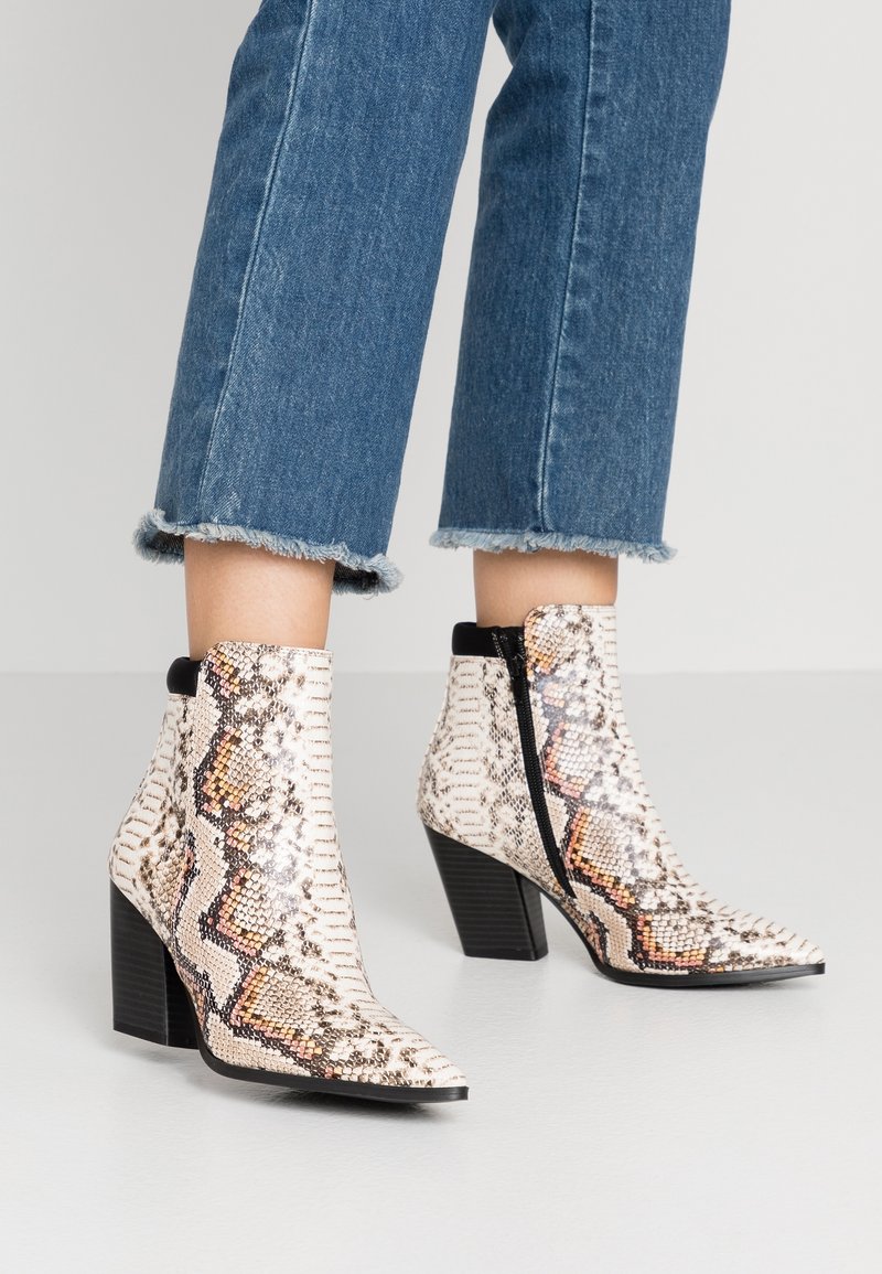 Botas tobilleras con estampado de piel de serpiente en blanco y beige, con punta afilada y tacón cuadrado, que cuentan con un cierre lateral para facilitar su uso.