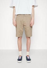 Shorts en coton beige à coupe droite, avec poches avant et fermeture par bouton, assortis à des baskets bleu marine et vertes, portés avec des chaussettes claires.