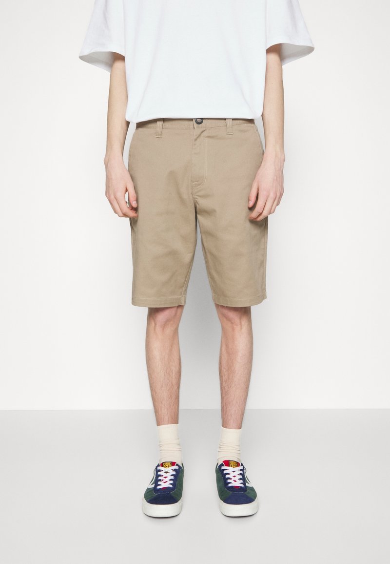 Shorts en coton beige à coupe droite, avec poches avant et fermeture par bouton, assortis à des baskets bleu marine et vertes, portés avec des chaussettes claires.