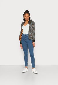 Casaco bomber floral com forro xadrez preto e laranja, sobre uma blusa verde-clara em V, combinado com jeans azul de cintura alta e ténis brancos.