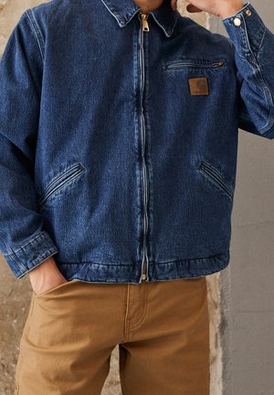 Homme portant une veste en jean bleue zippée et un pantalon marron, une main dans la poche et l'autre près du visage.