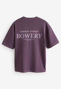 Violeta kokvilnas T-krekls ar īsām piedurknēm, apaļu kakla izgriezumu un priekšējo tekstu dizainā ar "BOWERY" un "23 WEST STREET" kontrastējošās krāsās.