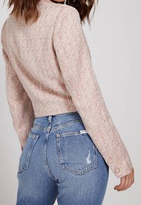 Giacca corta in tweed rosa con motivo strutturato, caratterizzata da maniche lunghe e bottoni decorativi, abbinata a jeans di denim a vita alta.