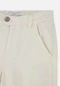Pantaloni in cotone beige con cintura elastica, tasche anteriori, chiusura con bottone e dettagli di trama sottile. Etichetta del marchio visibile all'interno.