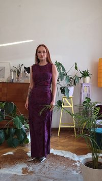 Vrouw in een mouwloze paarse jurk staat in een met planten gevulde woonkamer met houten meubels en een koeienhuiden kleed.