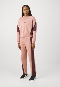 Roza in maroon trening obleka z dolgimi rokavi in prilegajočimi hlačami, izdelana iz mehkega materiala. Vsebuje Adidasovo logo na prsih in kontrastne stranske panele.