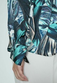 Blusa de manga larga con un vibrante patrón de hojas en azul y negro, con un puño abotonado y una textura de tela suave. Cortada a la cintura.