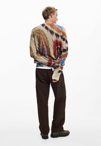 Maglione lavorato a maglia in beige con motivi astratti multicolori in rosso, blu e verde; abbinato a pantaloni larghi marrone scuro e scarpe robuste.