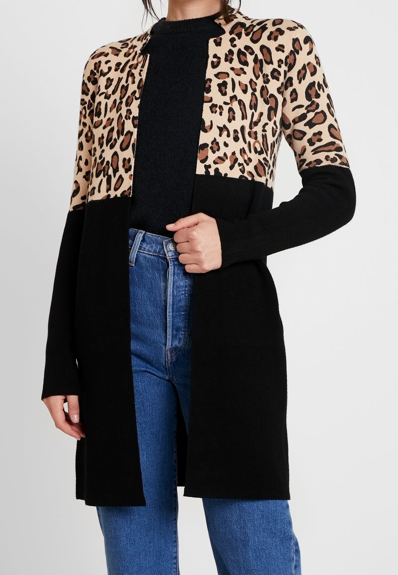 Femme portant un long cardigan noir avec une partie supérieure à imprimé léopard, un pull noir et un jean bleu taille haute, la main touchant le devant du cardigan.