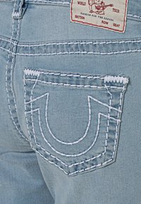 Světle modré džíny z denimu s výrazným bílým prošíváním na kapse a štítkem s červeným nápisem a grafikou na pásku.