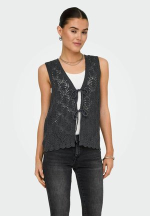 Grijze gehaakte vest met een golvende zoom, V-hals en sluiting met voorbind; gedragen over een witte top en gecombineerd met donkere jeans.