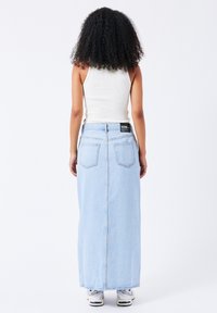 Falda maxi de mezclilla azul claro con corte recto, dos bolsillos traseros y etiqueta visible en la cintura. Combinada con una camiseta sin mangas blanca y zapatillas.