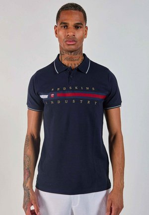Homme avec des tatouages au cou et au bras portant une chemise polo Redskins Industry bleu marine avec des rayures rouges, blanches et bleues, se tenant devant un fond neutre.