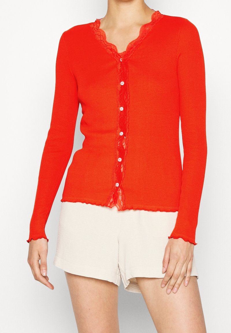 Cardigan rouge à côtes avec une bordure en dentelle le long du col et fermeture à boutons sur le devant. Manches longues et détail en bordure festonnée.