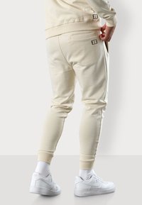 Beige Jogginghose mit Rippbündchen und zwei seitlichen Taschen. Mit einem Design-Tag auf der Rückseite. Kombiniert mit weißen Sneakers und Socken.