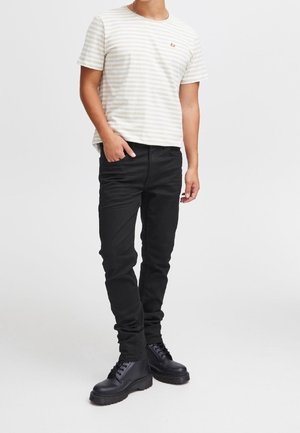 Mand iført en lysebeige og hvid stribet kortærmet t-shirt, sorte tætsiddende bukser og sorte snørebåndsstøvler, stående med den ene hånd i lommen.