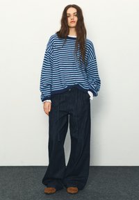 Modrý a námornícky pruhovaný nadmerný sveter, voľné tmavé džínsové nohavice, hnedé mokasíny. Mäkká textúra, pohodlný strih, ležérny štýl.