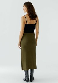 Schwarzes Tanktop mit dünnen Trägern, kombiniert mit einem olivgrünen gestrickten Midikleid. Das Model trägt hohe schwarze Stiefel mit Absatz. Insgesamt glatte Texturen.