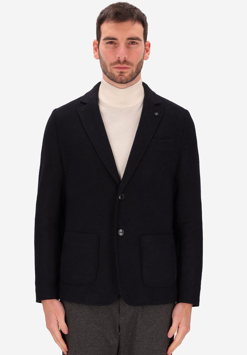 Uomo che indossa un blazer nero sopra un maglione a collo alto crema e pantaloni grigio scuro, in piedi contro uno sfondo grigio chiaro, rivolto in avanti.