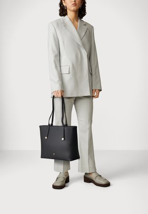 Vrouw in lichtgrijs maatpak en beige loafers, die een grote zwarte leren shopper met lange hengsels vasthoudt.