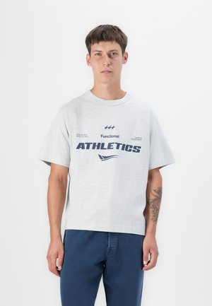 Grå bomulds-t-shirt med korte ærmer, der har marineblå tekst "Functional ATHLETICS" og dekorative grafik på fronten. Båret med blå bukser.