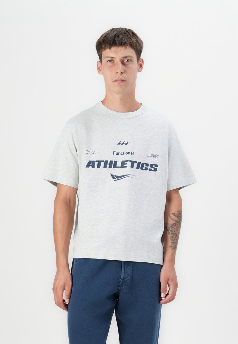 Graues Baumwoll-T-Shirt mit kurzen Ärmeln, das auf der Vorderseite den navyblauen Text "Functional ATHLETICS" und dekorative Grafiken zeigt. Getragen mit blauen Hosen.