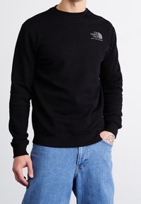 Felpa nera in cotone con scollatura a girocollo, polsini e orlo a coste, con un piccolo logo grigio sul lato sinistro del petto. Indossata con jeans azzurri chiari.