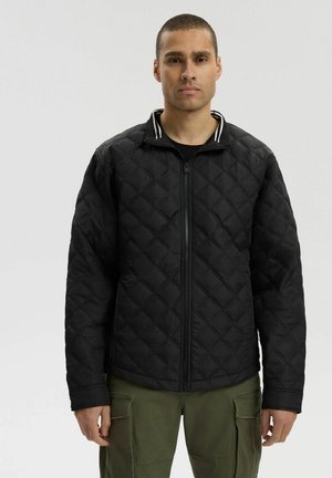 Mężczyzna ubrany w czarną pikowaną kurtkę zapinaną na zamek z detalem w postaci pasków na kołnierzu oraz oliwkowe spodnie cargo, stojący na tle jasnego, jednolitego tła.